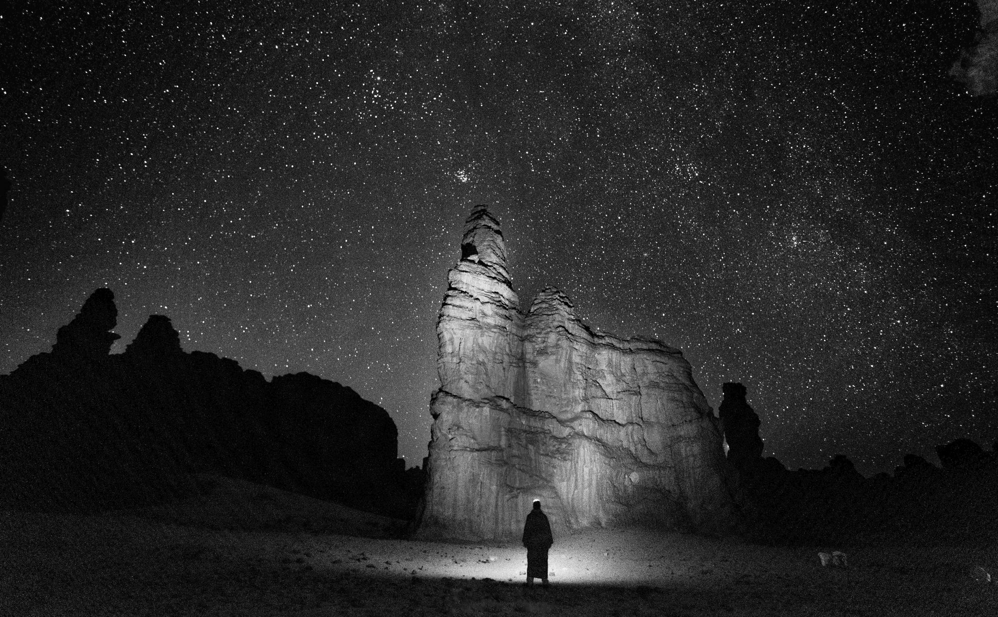 Nuit dans le Tassili n'Ajjer : une silhouette au pied d'un grand rocher éclairé à la lampe, sous un ciel plein d'étoiles.